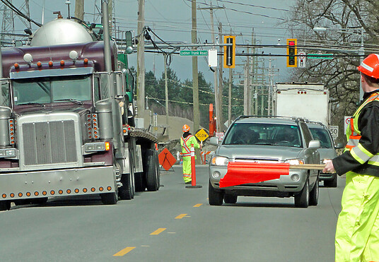 Busy road showing cars driving towards camera. A semi-truck is parked on the left side of the road and a worker, dressed in hi-vis, waves red flags to guide traffic.