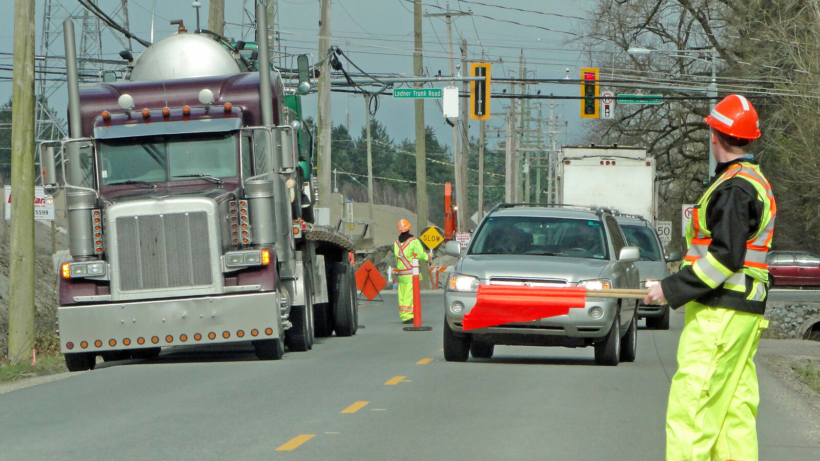 Busy road showing cars driving towards camera. A semi-truck is parked on the left side of the road and a worker, dressed in hi-vis, waves red flags to guide traffic.