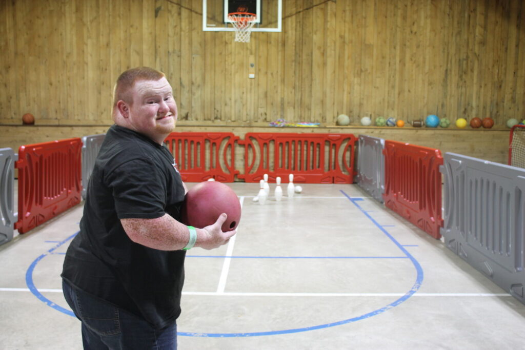 Alternating red and silver school barricades lined up to create an indoor bowling lane inside of a gym. Closest to the camera is a man with down syndrome smiling and holding a bowling ball.