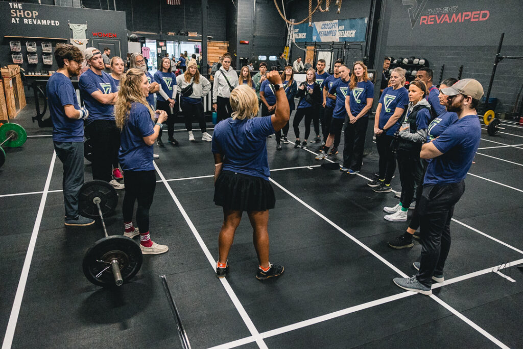 Crossfit athletes gather in matching competition t-shirts before beginning their tournament. They stand in the middle of the gym, lined with sports barricades.