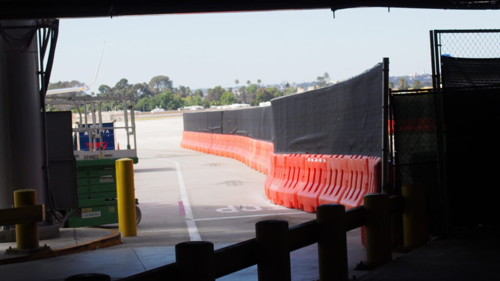 Orange OTW barricades with fencing and privacy screens line the perimeter of a construction site on the right side of the photo. Parts of an airport can be seen to the left and beyond.