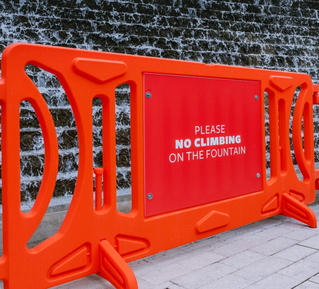 Bright orange pedestrian barricade on stone backdrop. Orange signage mounted to barricade reads: "Please no climbing on the fountain