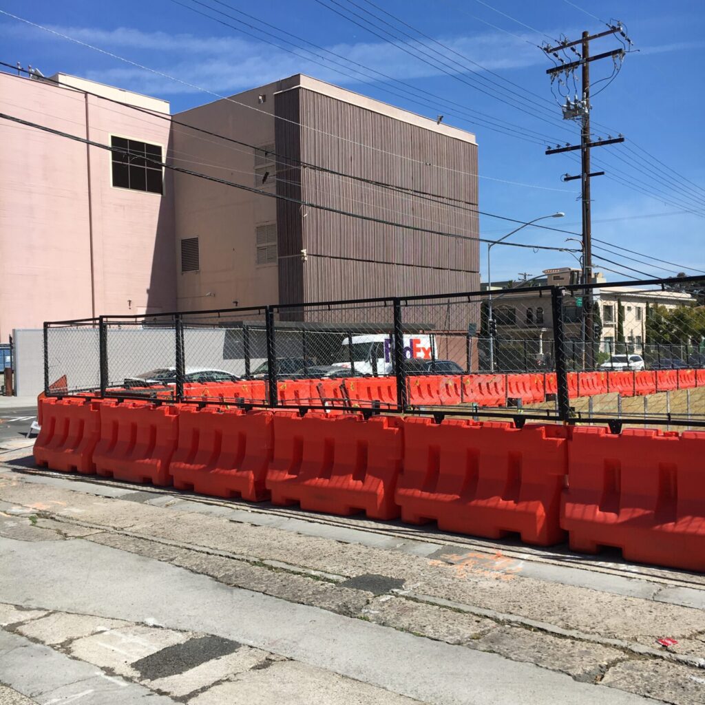 Orange OTW barricades with attached chain-link fencing surround a construction zone site in front of a brown building