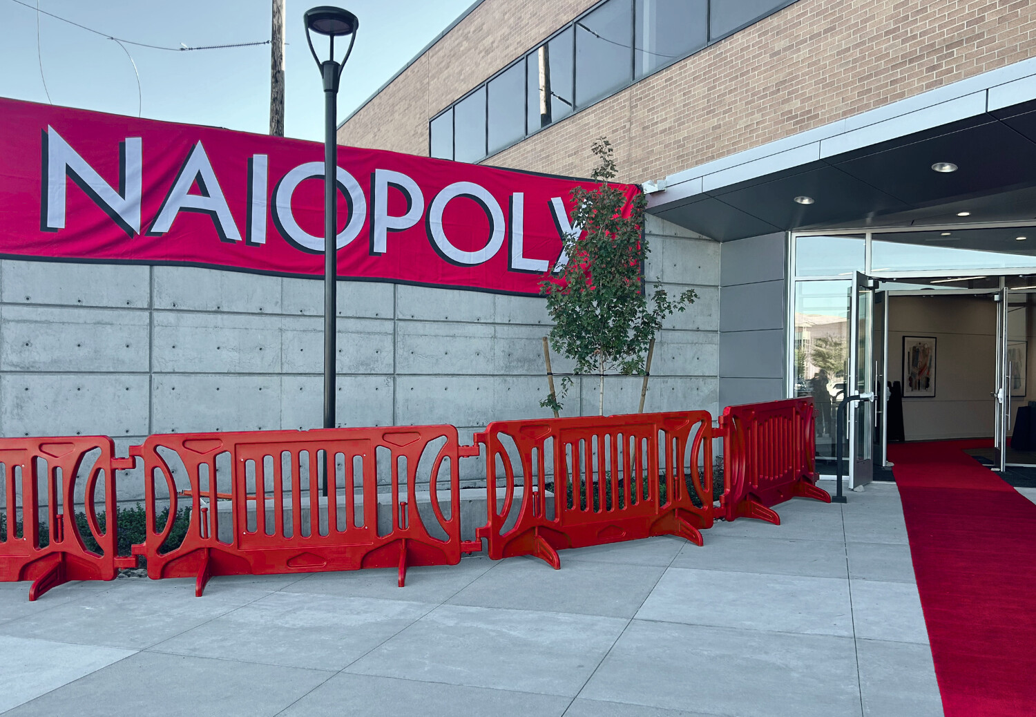 A red banner on a beige building spells NAIOPOLY, while red crowd control barricades create a line to the door underneath