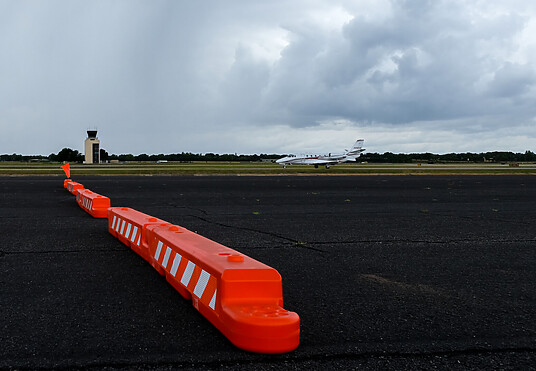 A line of orange and white low profile airport barricades start near the front and angle diagonally back on black asphalt, with a moody, cloudy sky in the background
