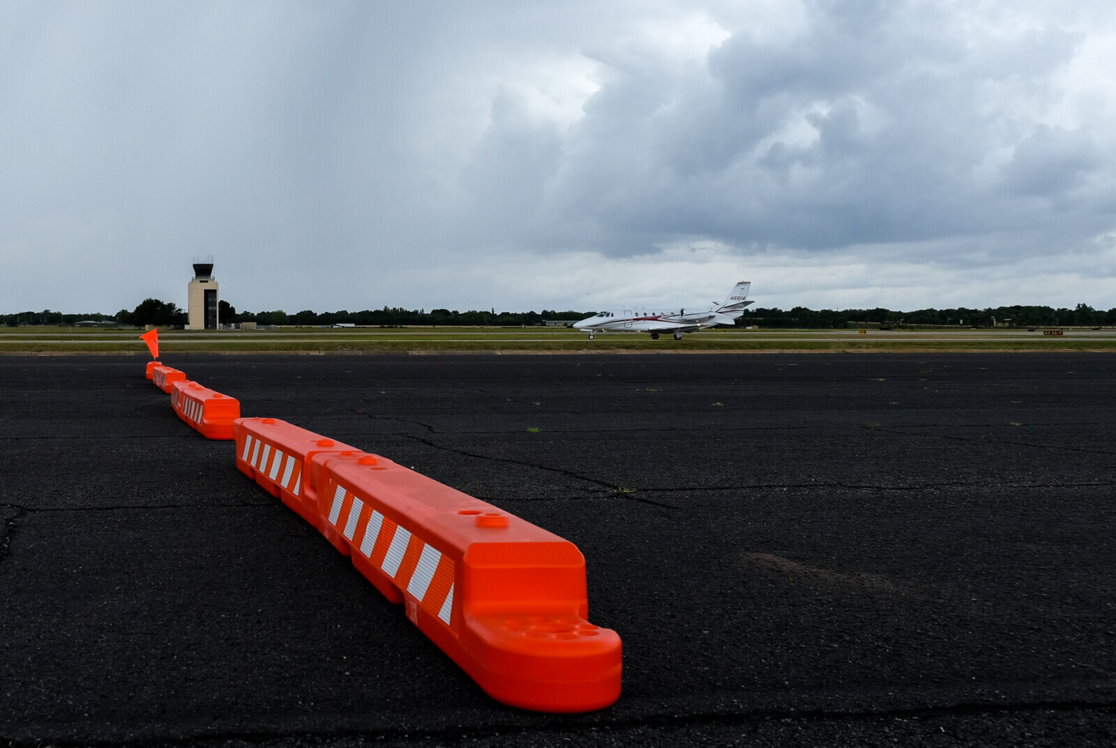 A line of orange and white low profile airport barricades start near the front and angle diagonally back on black asphalt, with a moody, cloudy sky in the background