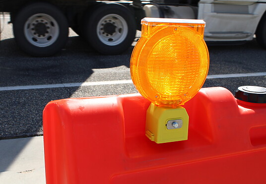An orange barricade hazard light is attached to a close up of an orange barricade, with the wheels of a semi truck in the background.