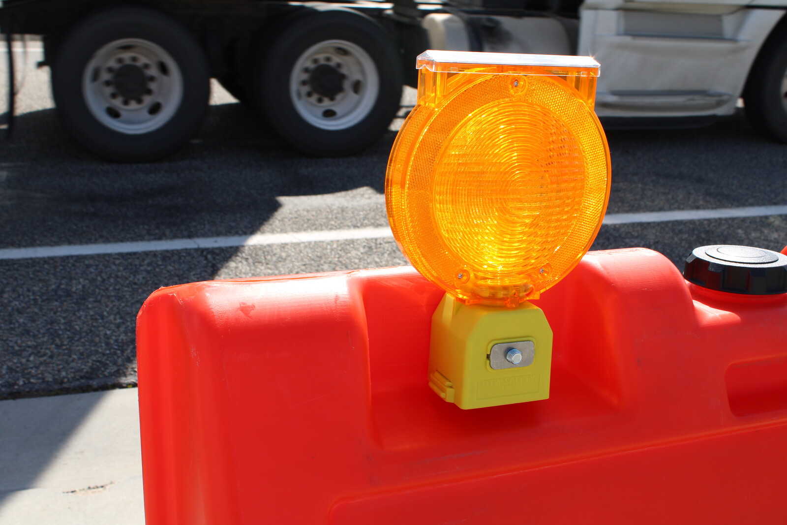 An orange barricade hazard light is attached to a close up of an orange barricade, with the wheels of a semi truck in the background.