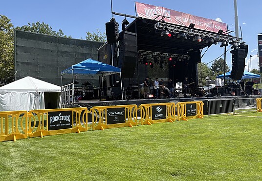 Yellow OTW Safety event barricades with black and white signage are set up in front of a large stage with a red awning.