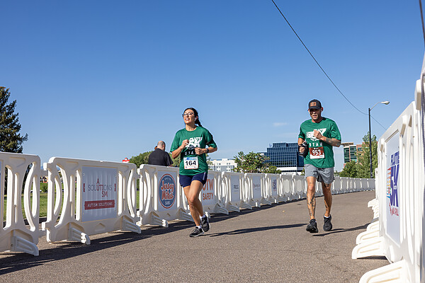 Two runners dressed in green run on a paved walkway alongside a line of connected white OTW event barricades