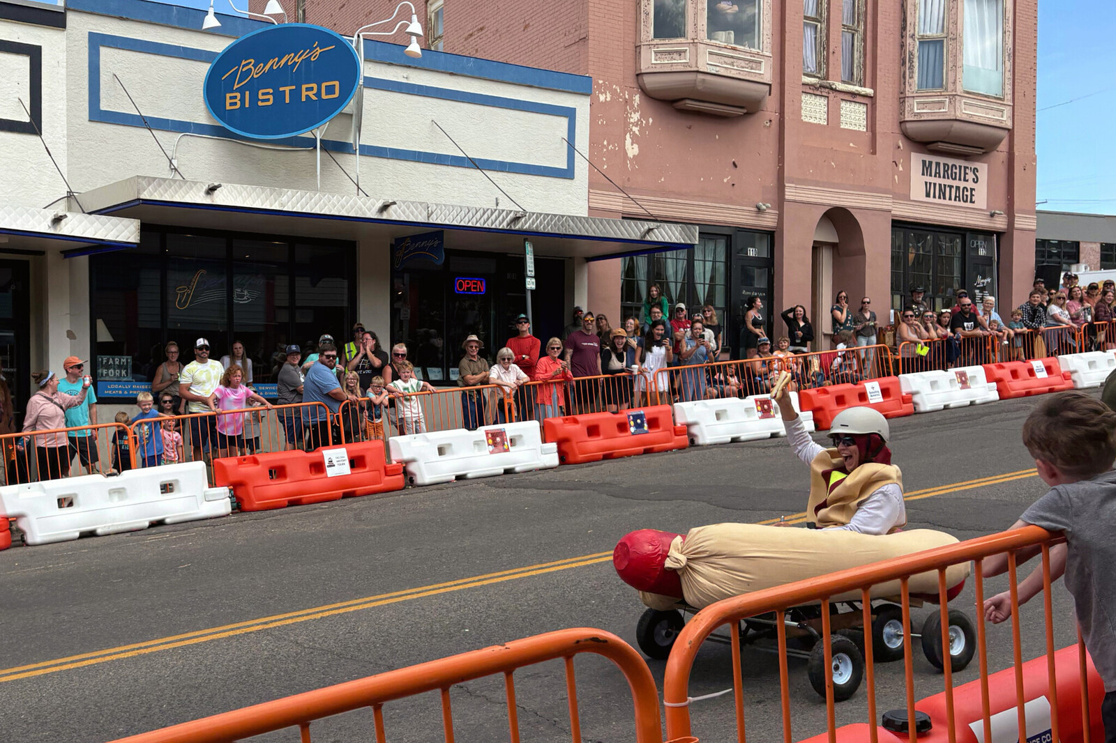 A hot dog shaped derby car is caught in motion racing down a track lined by white and orange go kart barricades