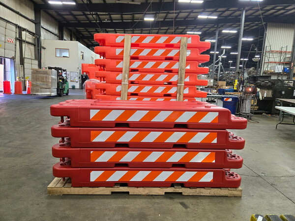 A pallet of custom red OTW airport barricades sits in front of a taller pallet of regular safety orange OTW airport barricades, highlighting the difference in color between the two.
