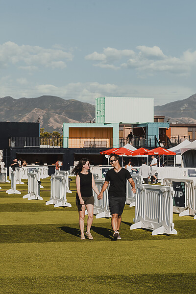 White pedestrian barricades are spaced out on a green field. Two people are walking and holding hands beside them. Umbrellas and cargo containers appear in the background.