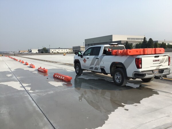 A white truck is parked on a runway, loaded with orange low-profile barricades. To the left, the same barrricades are set up in a line going away from the truck