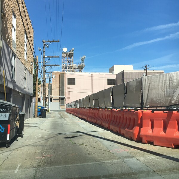 Tall buildings rise on the left, while orange construction barricades with added fencing panels line the street on the right side of the photo