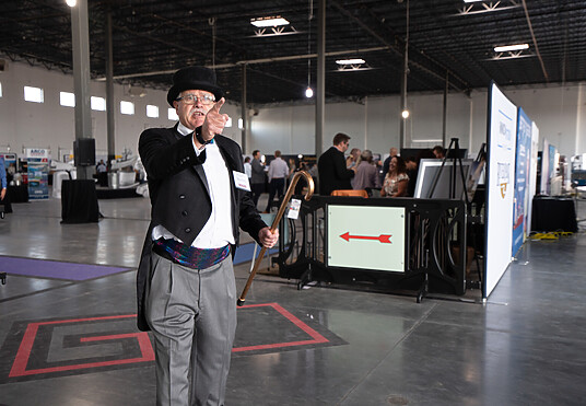 A man in suit tails and a balck top hat speaks to someone out of frame. Behind him, a black pedestrian barrier, the Billboard Barricade, is set up with custom signage on it to welcome people to the event Naiopoly.